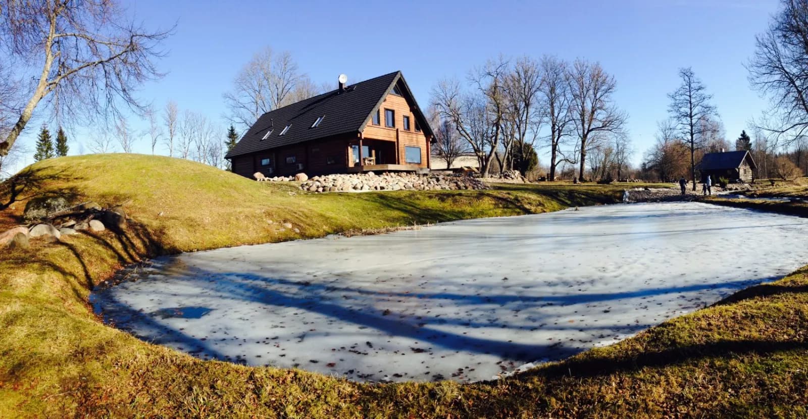 Tumedaks lasuuritud palkmaja tiigi ääres, viilkatusel (mitte kelpkatusel) kaks katuseakent, jäätunud tiik, pruunikas muru ja lehtedeta puud, lund maas ei ole. Töö tegi L.L. House.