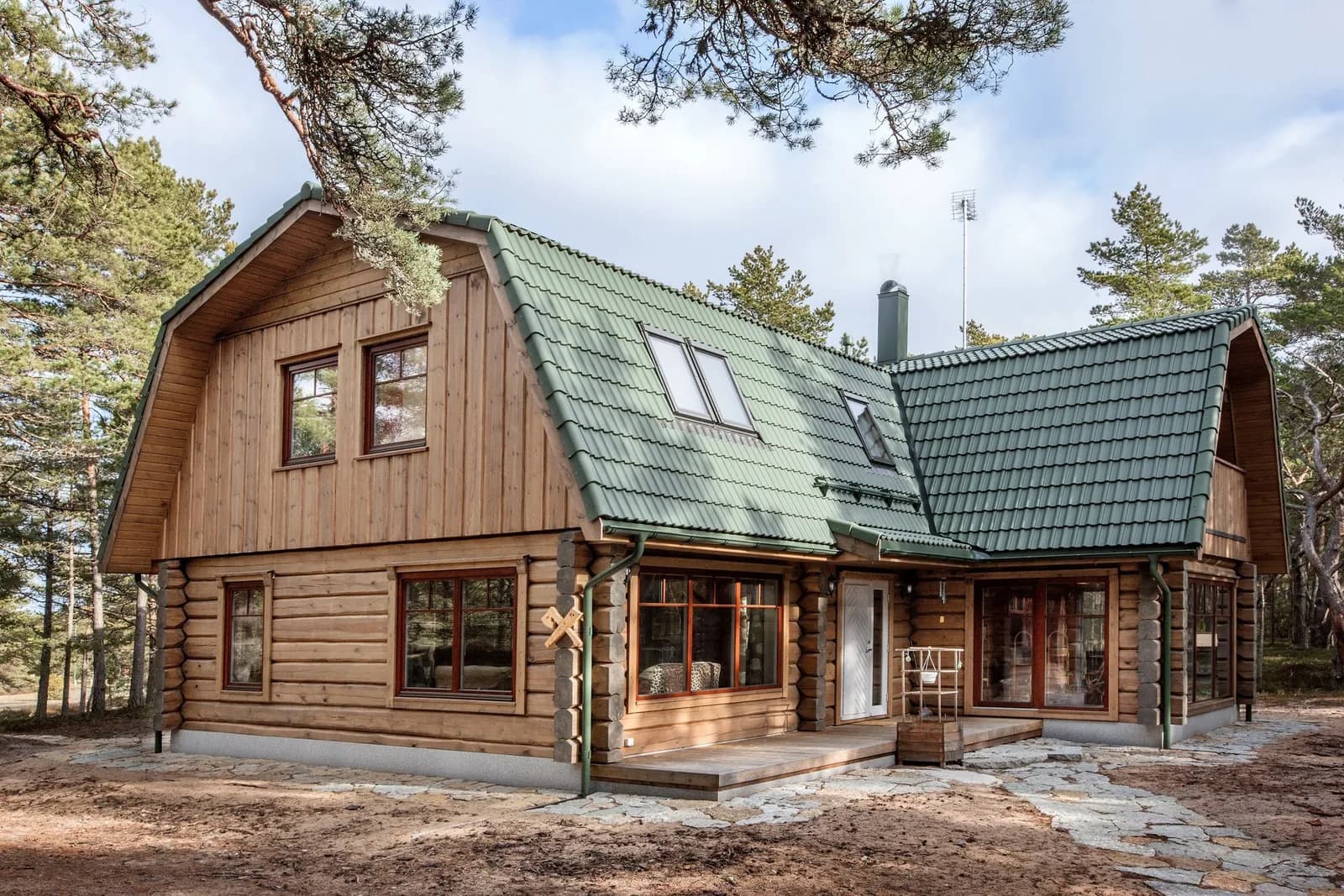 Kahekorruseline palkmaja tumerohelise kelpkatuse ja valgete aknaraamidega, liivasel-kivisel hoovialal männimetsas, selge taevaga. Töö tegi Eesti Käsitöö Palkmajad.